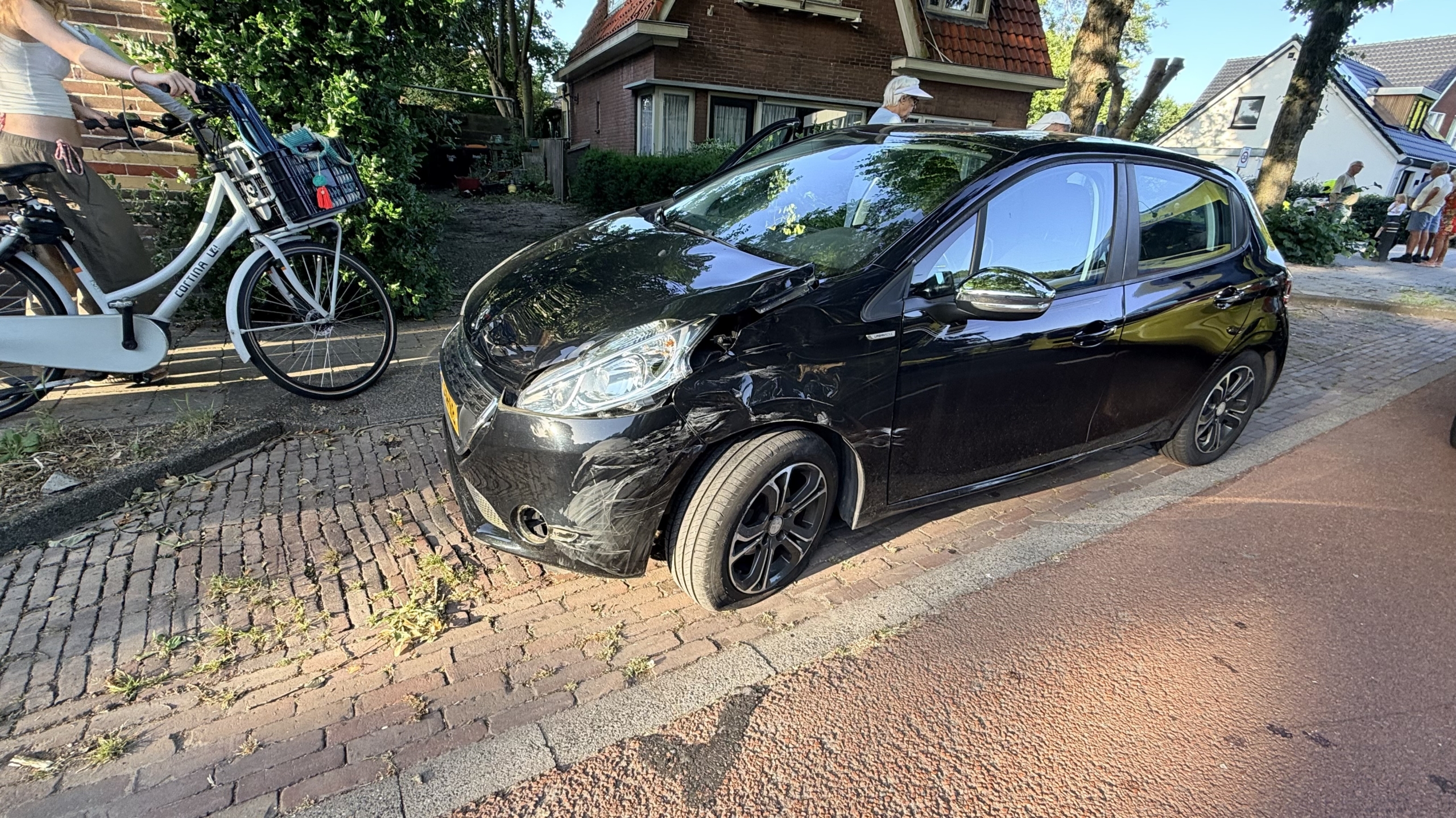 Omgevallen motorfiets op de stoep naast een ambulance en politieauto, met omstanders aan de kant van de weg.