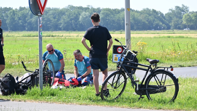 Twee mensen in sportkleding verlenen hulp aan een persoon naast een fiets, met een kijker en een verkeersbord op de achtergrond.