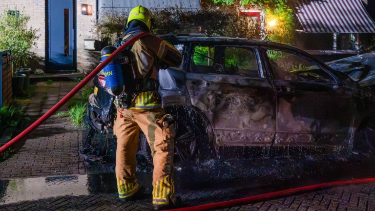 Brandweerlieden blussen een brandende auto op een straat 's nachts, omringd door rook en water.