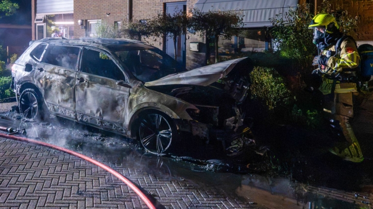 Brandweerlieden blussen een brandende auto op een straat 's nachts, omringd door rook en water.