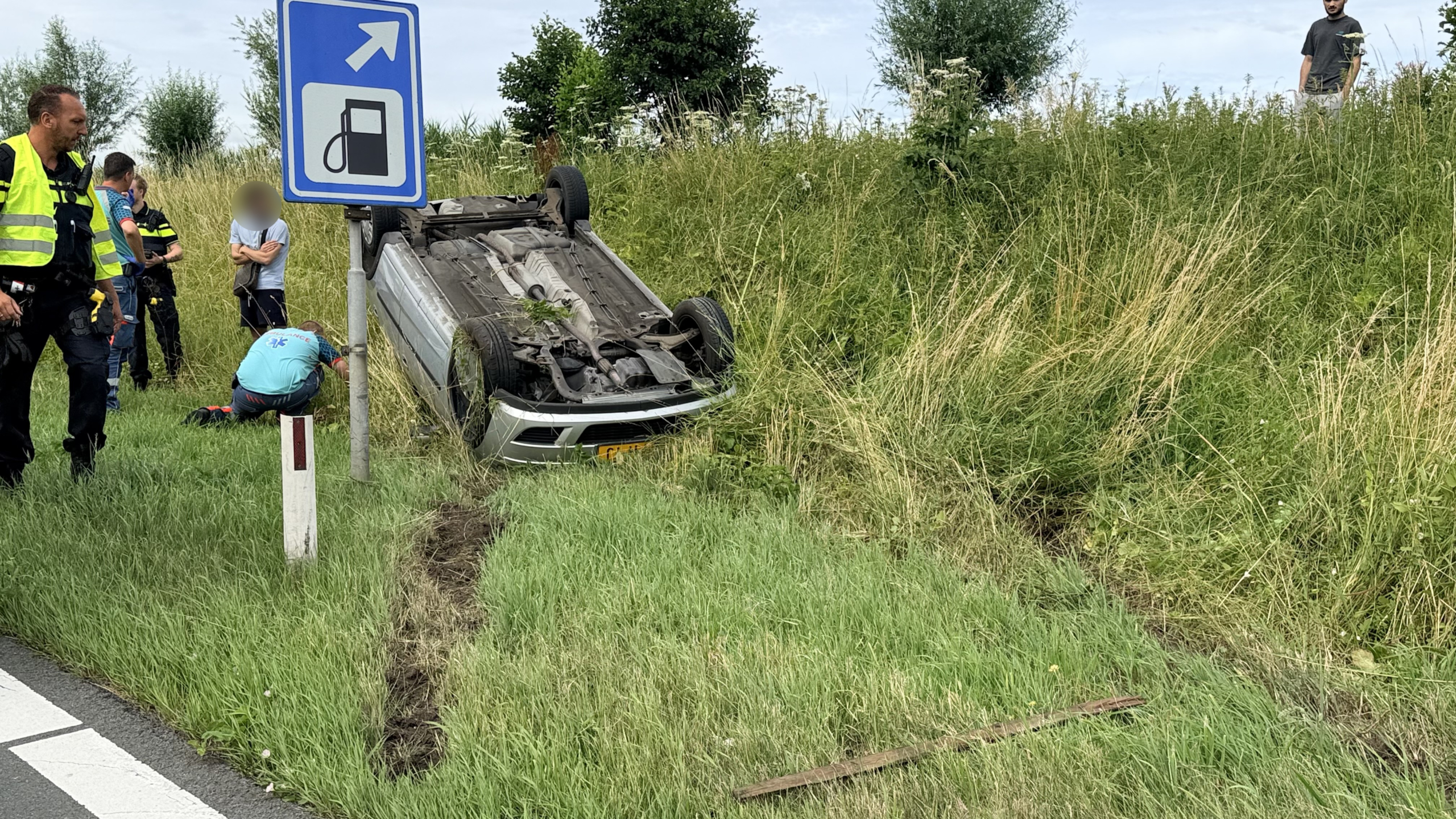 Auto ligt ondersteboven in de berm naast een weg, met politieagenten en een ambulance aanwezig.