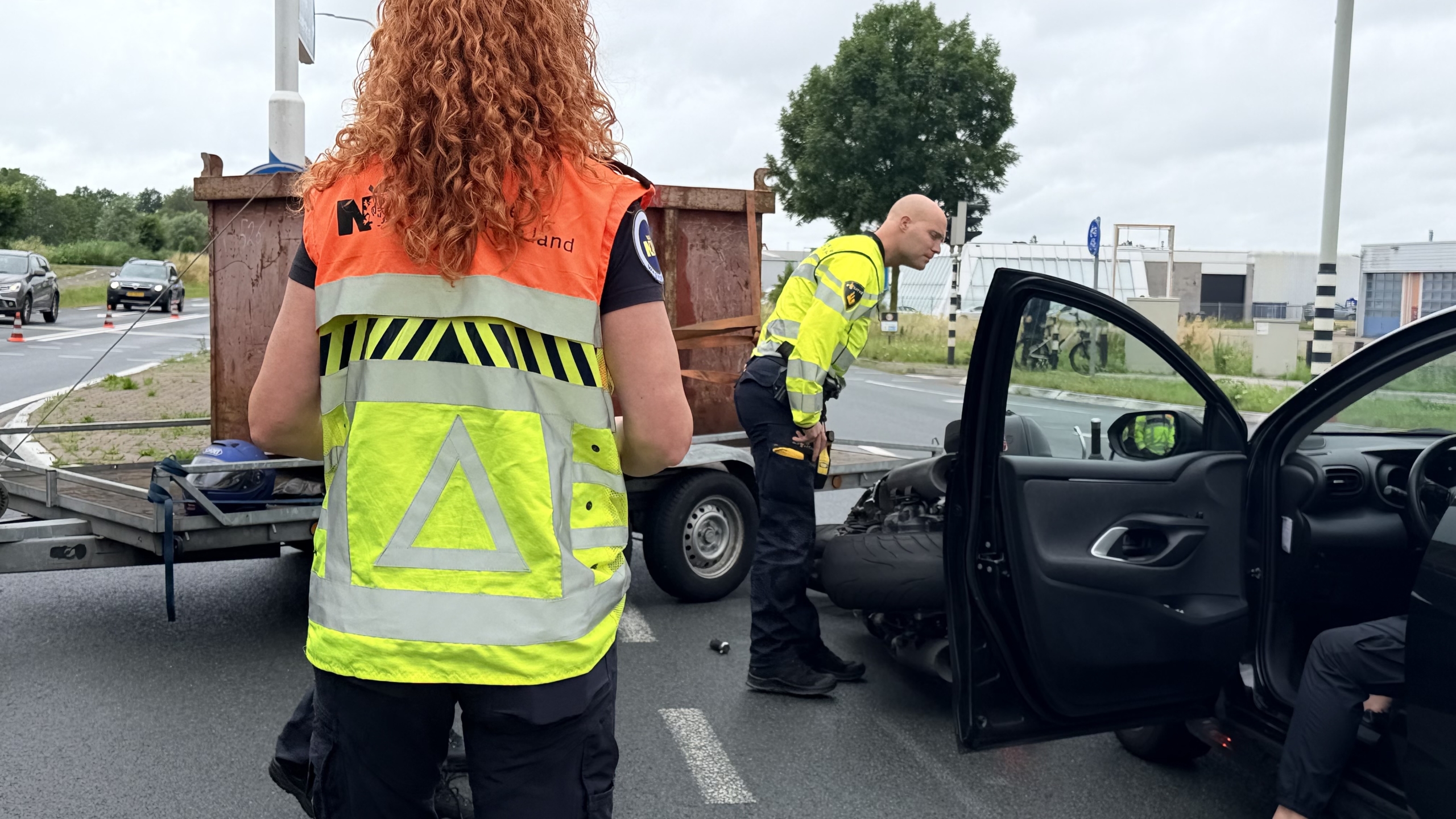 Omgevallen motorfiets op de grond naast een auto en aanhanger op een kruispunt, met verkeer en verkeerskegels op de achtergrond.