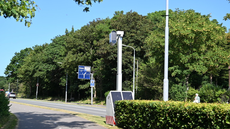 Snelheidscamera langs een weg met fietspad, verkeersborden en hoge bomen op een zonnige dag