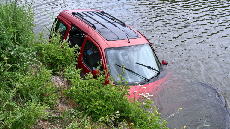Rode auto half onder water in een rivier, met de voorkant en voorruit zichtbaar naast een begroeide oever