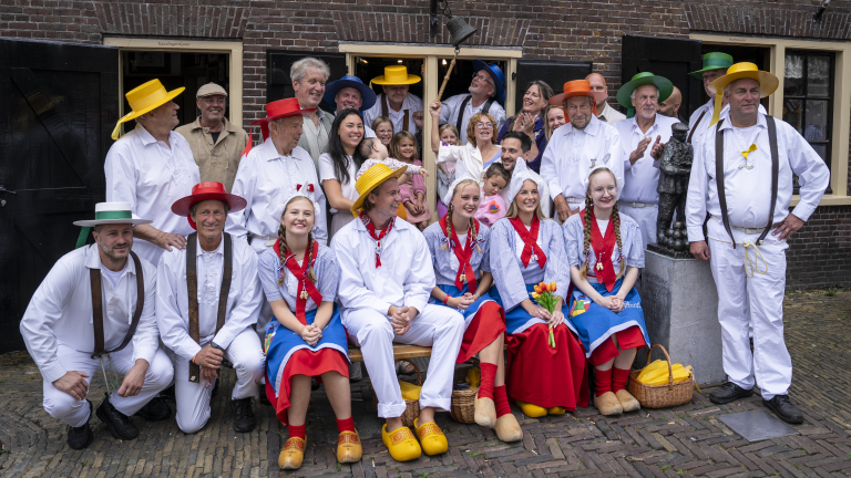 Groepsfoto van mensen in traditionele Nederlandse klederdracht met kleurrijke hoeden voor een bakstenen gebouw