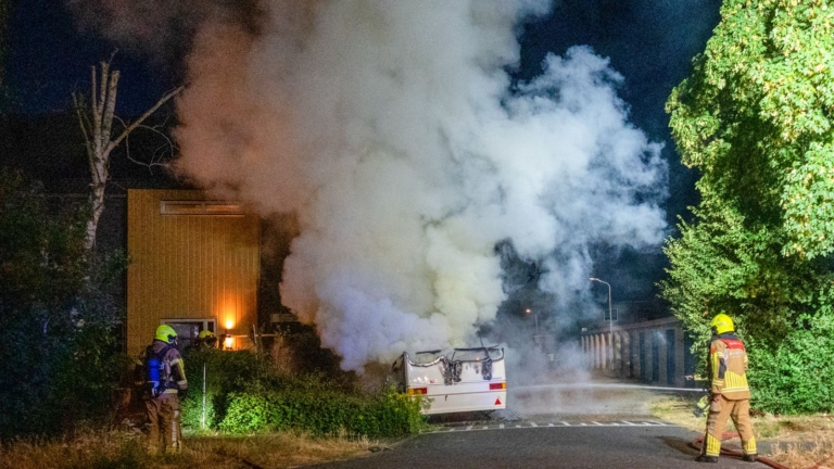 Brandende camper naast een gebouw in de nacht, met hoge vlammen en dikke rookpluim langs een rij garageboxen