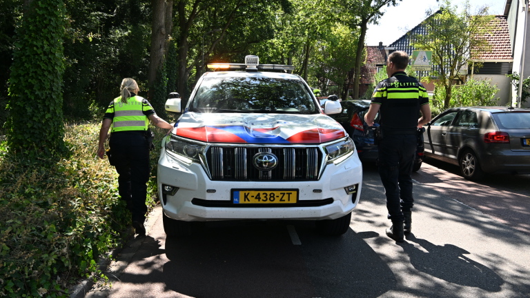 Twee politieagenten lopen langs een geparkeerde politieauto op een straat met bomen en woonhuizen