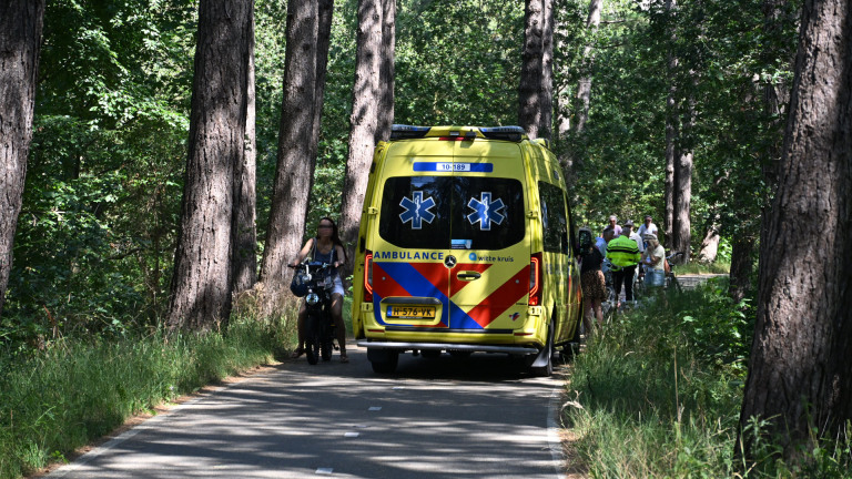 Gele ambulance op een bospad, met fietsers en omstanders eromheen tussen hoge bomen
