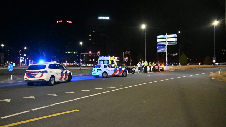 Politieauto en ambulance met zwaailichten bij nachtelijk ongeval; motorfiets en helm op de weg, agent regelt verkeer