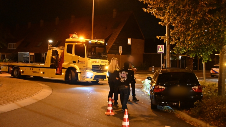 Zwarte auto met zware frontschade tegen een paal, ’s nachts; bestuurder stapt uit, met verkeerskegel op de weg