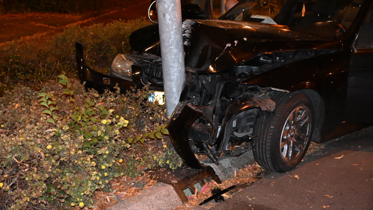 Zwarte auto met zware frontschade tegen een paal, ’s nachts; bestuurder stapt uit, met verkeerskegel op de weg
