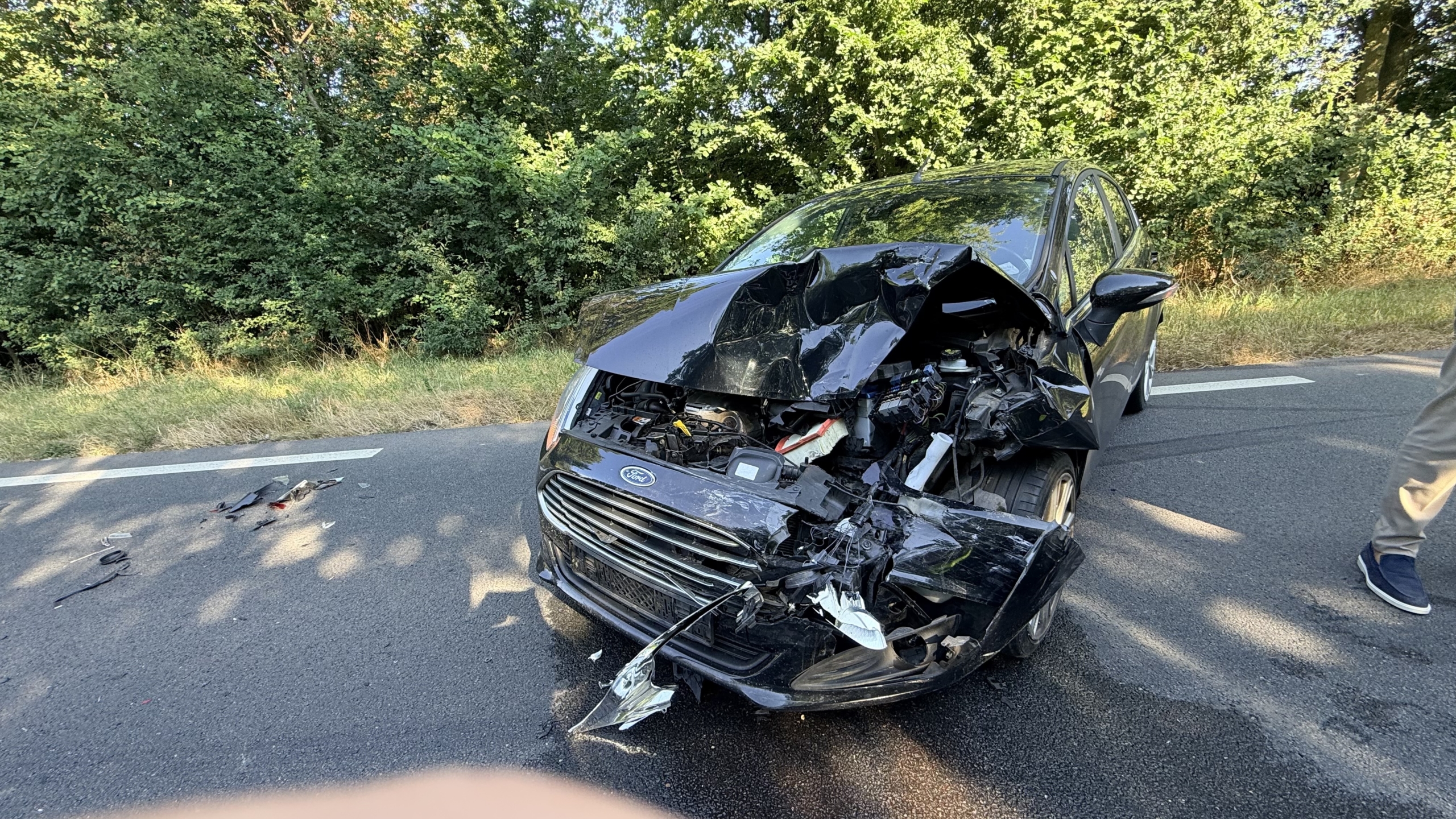 Ambulance en politie bij auto-ongeluk op bosrijke weg, met beschadigde zwarte auto en brokstukken op het asfalt