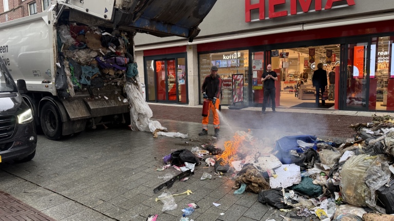 Vuilniswagen lost rokende afvalberg voor de HEMA, terwijl twee medewerkers blussen en omstanders bij de ingang staan