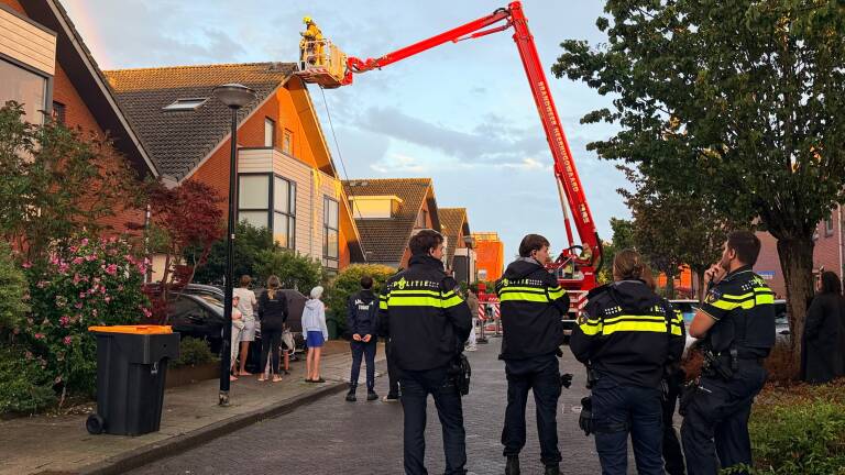 Politie en omwonenden kijken toe terwijl brandweer met hoogwerker een dak van een woonhuis inspecteert