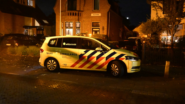 Politieagent telefoneert naast een politieauto op een donkere woonstraat, met een beschadigde paal op de voorgrond