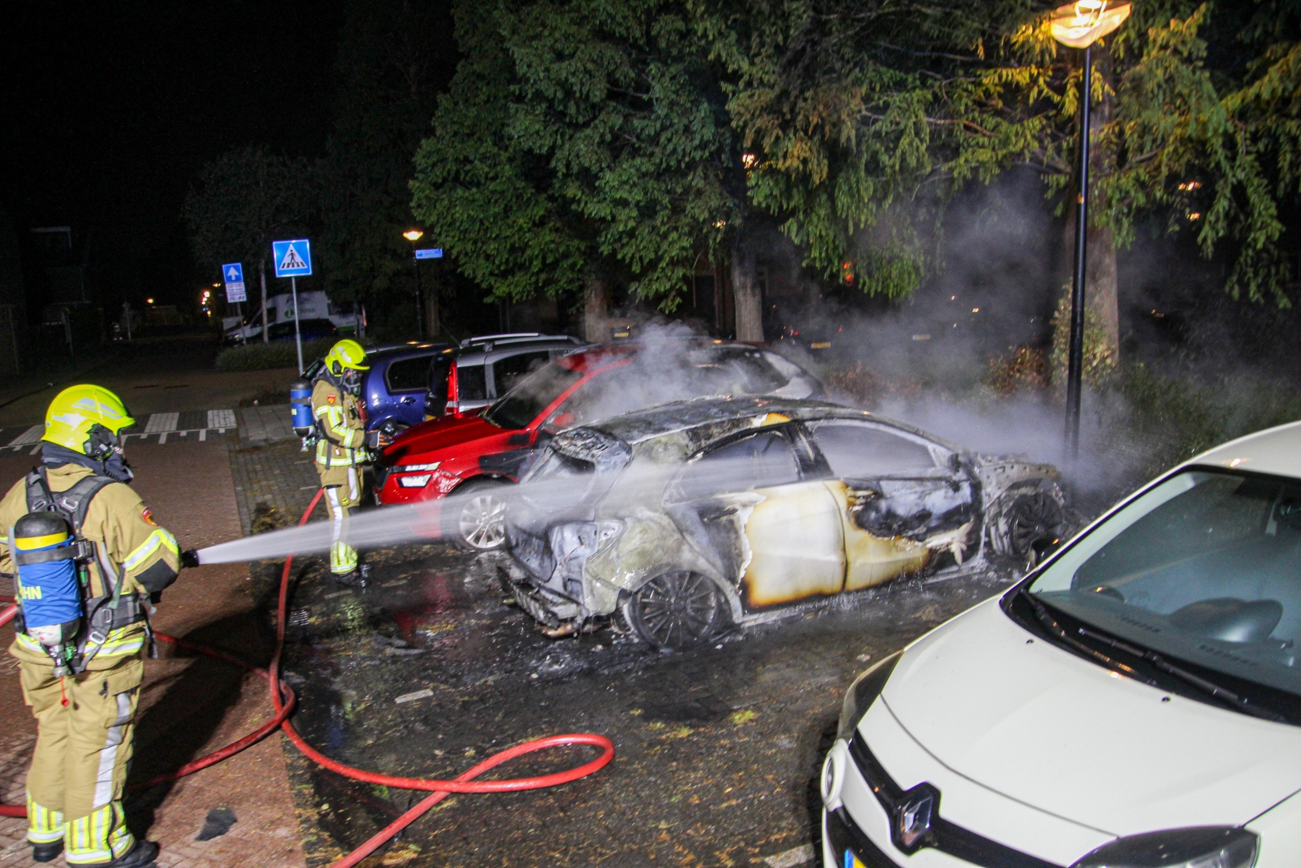 Brandweerlieden blussen brandende auto op parkeerplaats, met veel rook tussen geparkeerde auto’s in het donker