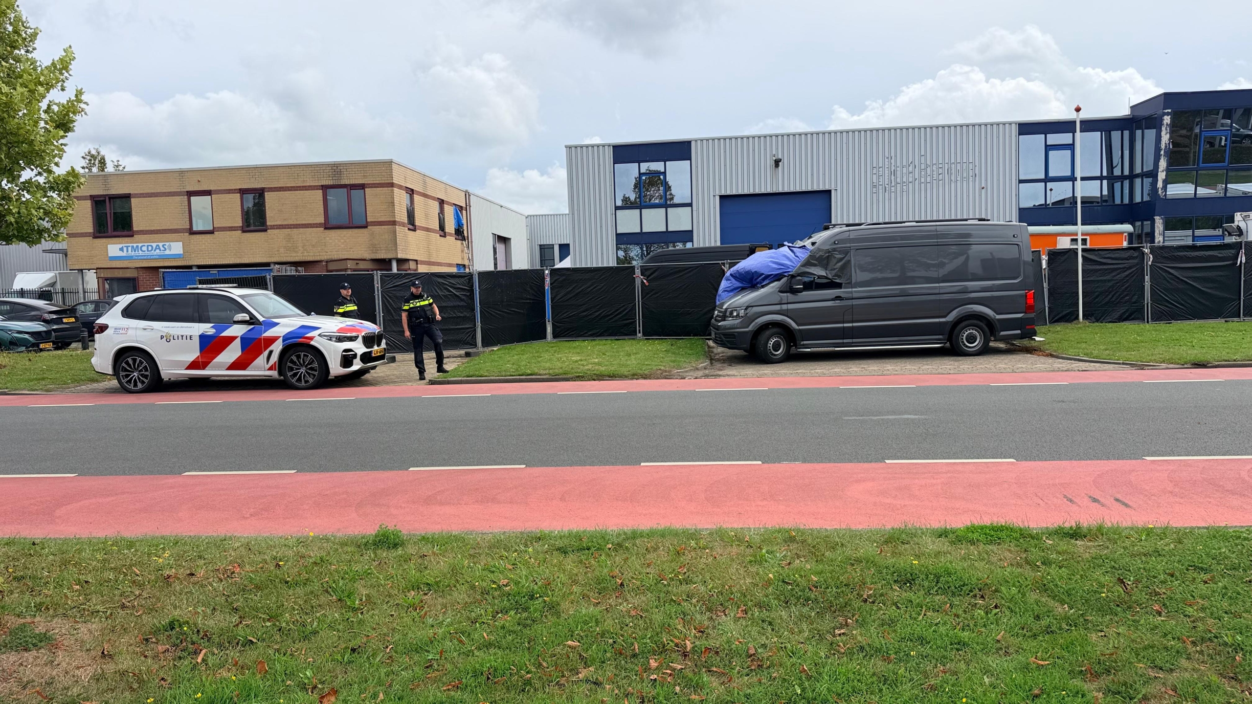 Politieagenten bij een grijze bestelbus met blauw zeil op een bedrijventerrein, met politieauto op de voorgrond