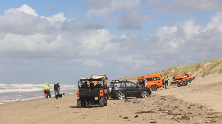 Groep hulpverleners op strand kijkt naar gekantelde zeilboot die is vastgelopen in de branding