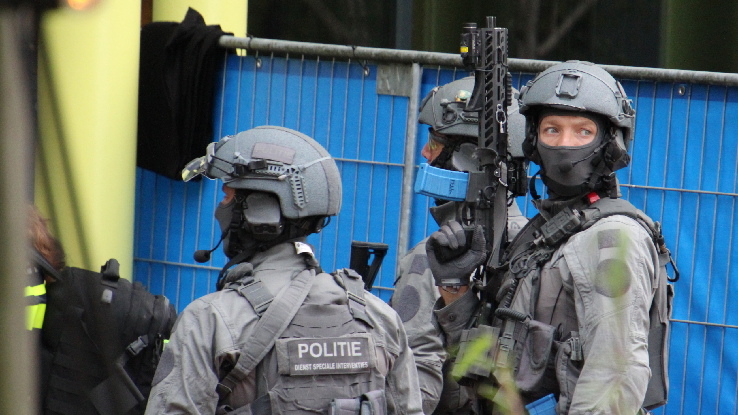 Zwaarbewapende politieagenten in grijze tactische uitrusting met helmen staan in groep bij een blauw hek