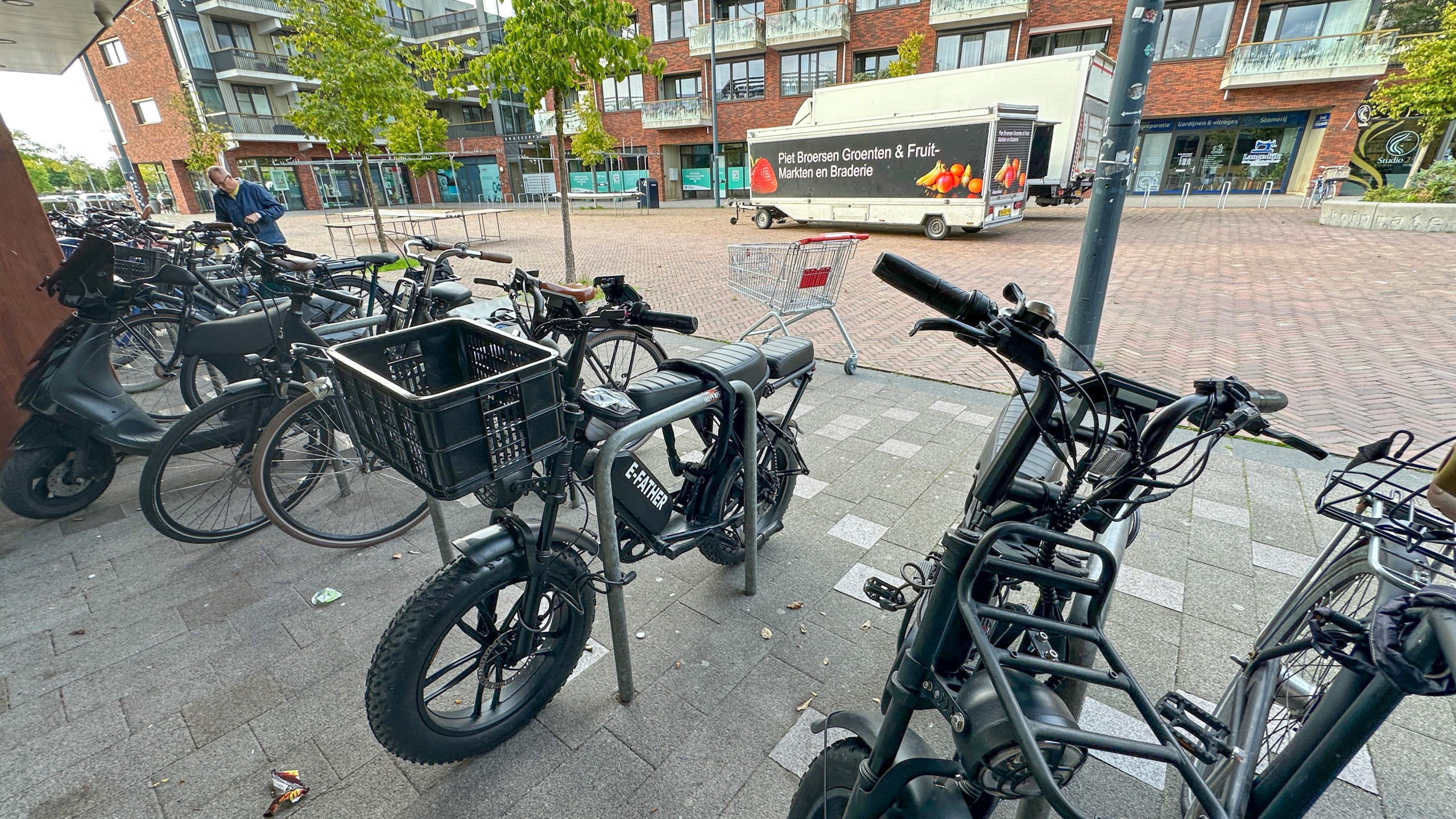 Zwarte e-fatbike met grote mand voorop geparkeerd bij fietsenrekken op een stoep, met flats en bomen op de achtergrond