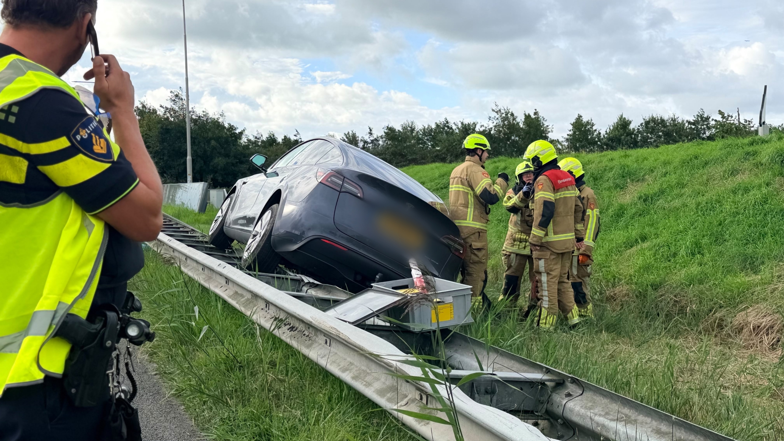 Grijze auto staat scheef op de vangrail langs een afrit, met brandweer en ambulance op de achtergrond