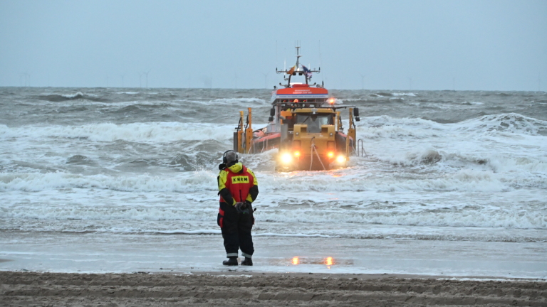 Reddingsboot vaart door ruwe zee naar strand terwijl een KNRM-hulpverlener toekijkt