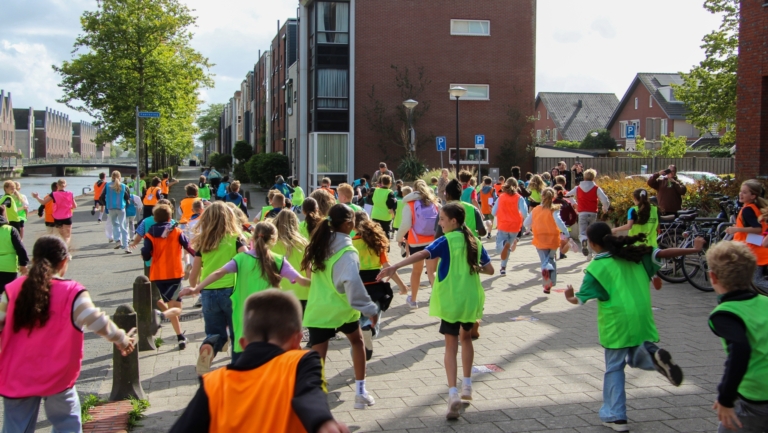 Grote groep kinderen in felgekleurde hesjes rent door een woonwijk langs een kanaal, tijdens een hardloopactiviteit