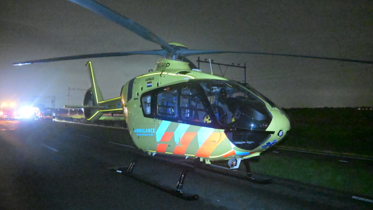 Ambulance met zwaailichten en hulpverleners langs een donkere weg, met een auto in de berm