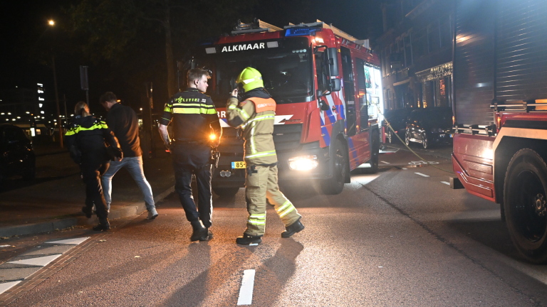 Brandweerlieden en politie op straat bij een hoekpand in de nacht, met brandweerwagen, slangen en fietsen op de voorgrond