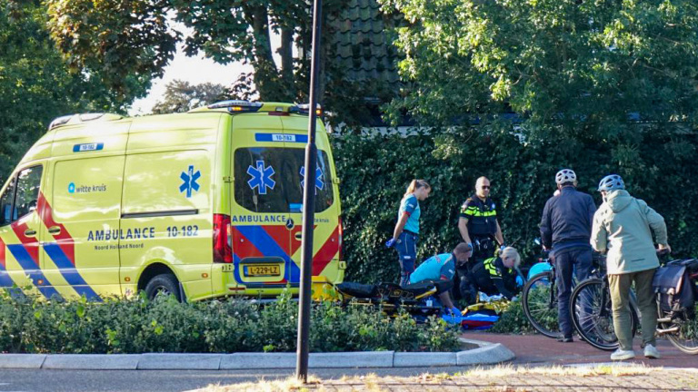 Ambulance geparkeerd langs de weg terwijl hulpverleners een slachtoffer behandelen; omstanders met fietsen kijken toe