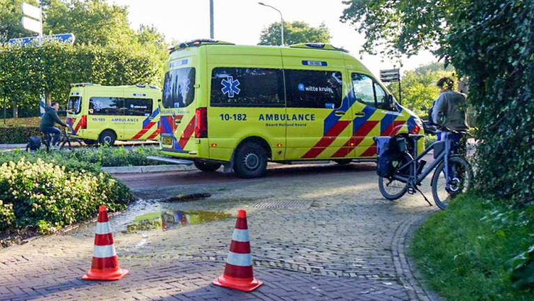 Ambulance geparkeerd langs de weg terwijl hulpverleners een slachtoffer behandelen; omstanders met fietsen kijken toe