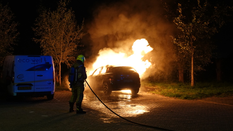 Brandweerman blust een brandende auto met grote vlammen en rook op een donkere weg naast een bestelbus