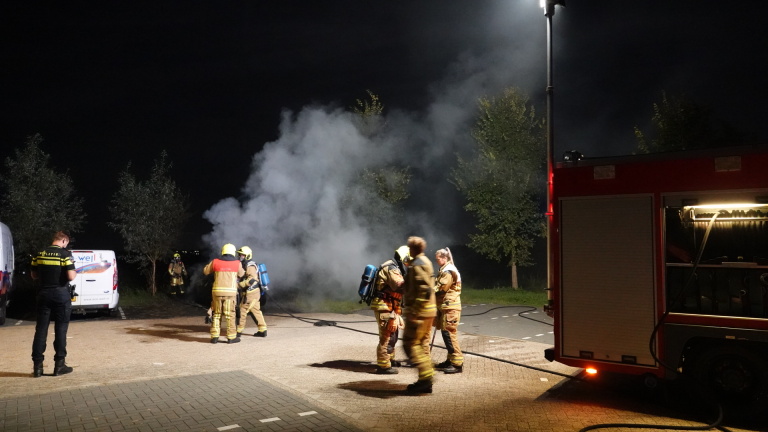 Brandweerman blust een brandende auto met grote vlammen en rook op een donkere weg naast een bestelbus