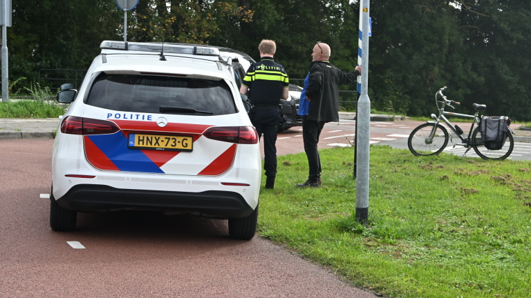 Gele ambulance met open zijdeur; politieagent staat ernaast langs een weg met groen en bordje Dijkweg