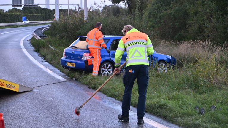 Blauwe auto met schade aan de zijkant staat deels in de berm naast een weg, omringd door gras en struiken
