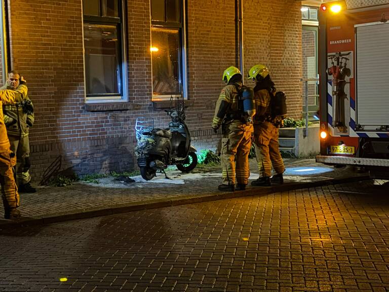 Brandweerlieden bij een bakstenen gebouw naast een brandweerwagen, inspecteren een uitgebrande scooter op straat