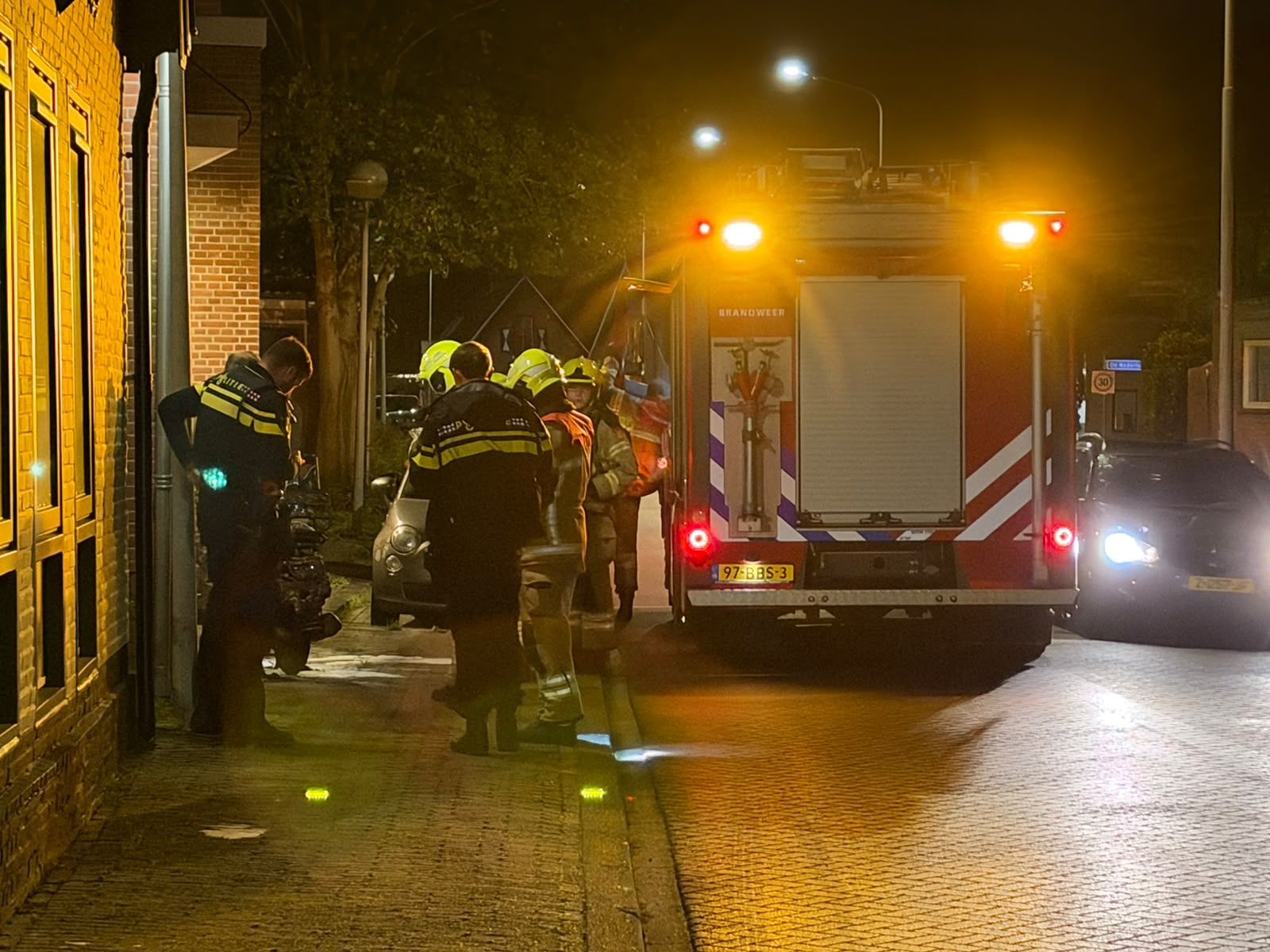 Brandweerlieden bij een bakstenen gebouw naast een brandweerwagen, inspecteren een uitgebrande scooter op straat