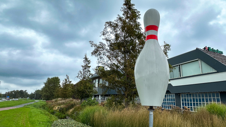 Grote bowlingkegel met rode strepen naast een gebouw met Heineken-logo, onder een bewolkte lucht