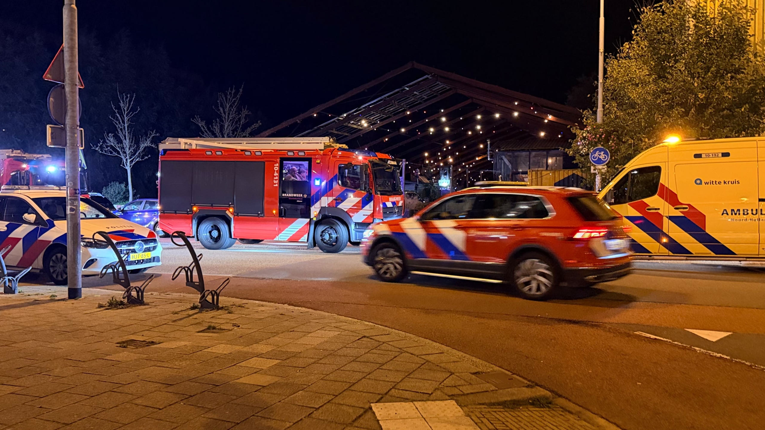 Brandweerlieden staan ’s avonds bij een deels uitgebrande loods met gebroken ruiten en een groot graffiti-paneel ervoor