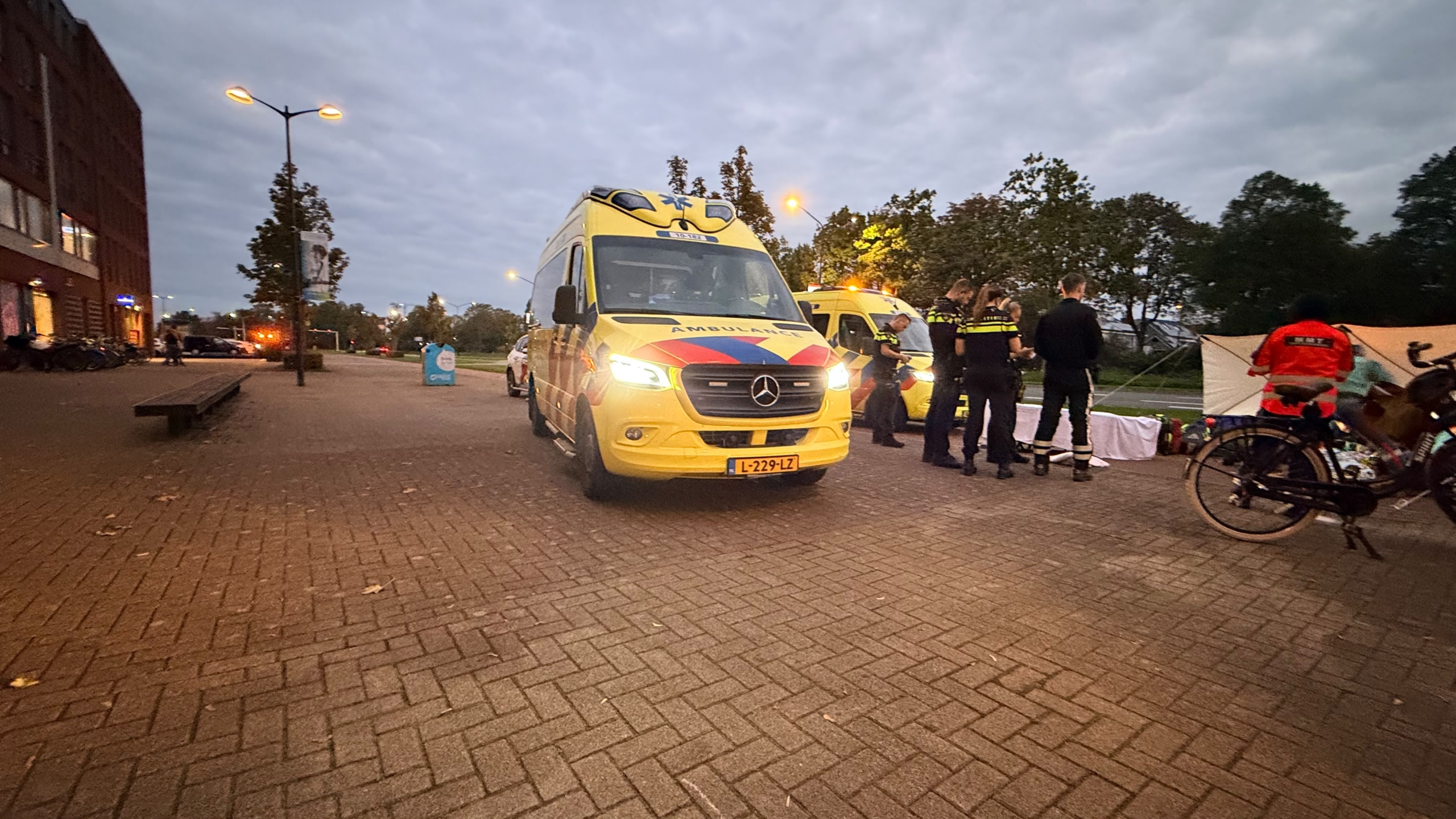 Twee ambulances met hulpverleners die iemand behandelen bij een wit afschermdoek langs de weg in de schemering