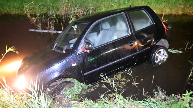 Zwarte auto deels in een sloot beland langs een grasberm, met modder op de carrosserie en koplampen aan