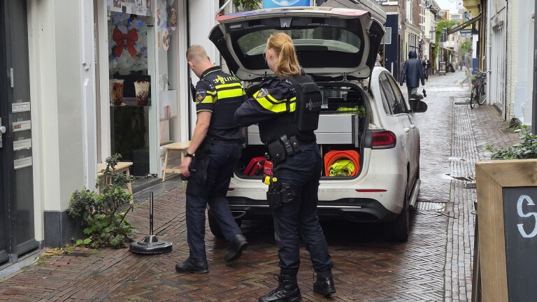 Twee politieagenten staan bij een politieauto met open achterklep in een smalle straat bij een winkelpand