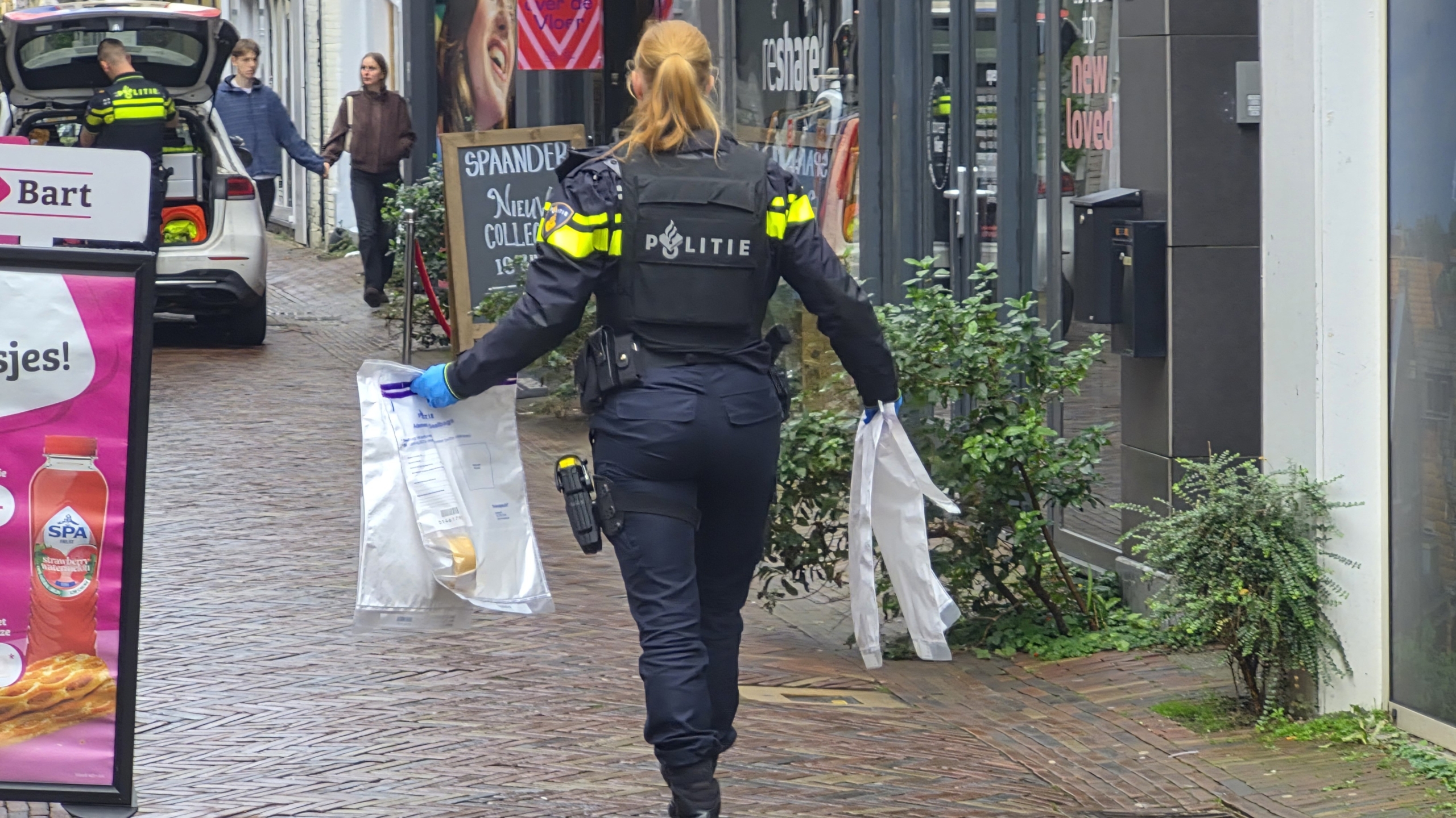 Twee politieagenten staan bij een politieauto met open achterklep in een smalle straat bij een winkelpand