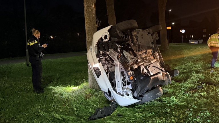 Omgeslagen auto tegen een boom op een grasveld bij nacht, met politieagent en hulpverlener in de buurt