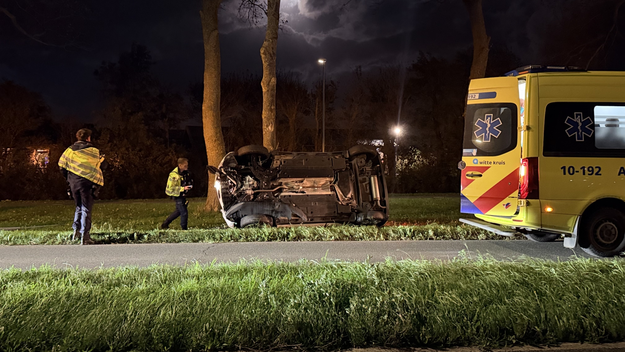 Omgeslagen auto tegen een boom op een grasveld bij nacht, met politieagent en hulpverlener in de buurt