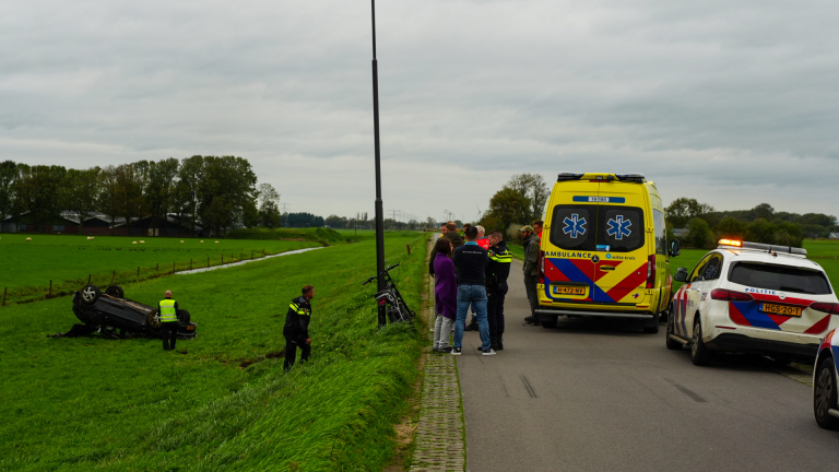 Omgeslagen auto op een grashelling, met ambulances en hulpdiensten op de weg erboven